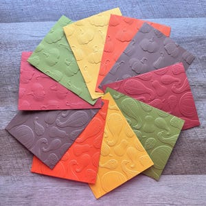 May include: A collection of colorful, textured paper squares arranged in a circular pattern. Each square features an embossed design of pumpkins and gourds in shades of orange, yellow, green, and burgundy. The overall theme is autumn and harvest.