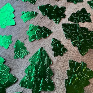 May include: A collection of shiny, emerald green Christmas tree-shaped embellishments. The trees vary in size and feature embossed patterns, including swirls, snowflakes, and geometric designs. They are scattered on a wooden surface.