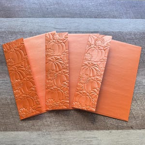 May include: A set of orange stationery items including two tall, narrow bookmarks and two rectangular envelopes, all featuring a raised, embossed pattern of pumpkins and sunflowers across the surface of the paper.