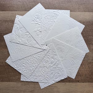 May include: A collection of white, textured cards arranged in a circular pattern. Each card features a different embossed floral or botanical design, including roses, leaves, and abstract patterns. The cards are displayed on a wooden surface.