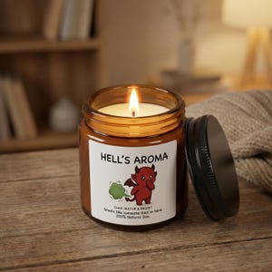 Pode incluir: Uma vela acesa em um frasco de vidro marrom com tampa preta. O rótulo diz "HELL'S AROMA" com uma ilustração de diabo de desenho animado. O aroma é "SULFUR & REGRET".