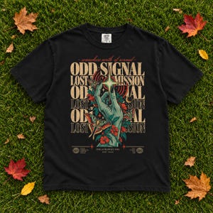 May include: A black short-sleeved t-shirt featuring a graphic print of a hand reaching towards a star, surrounded by moths and flowers. The text reads: somewhere north of normal, ODD SIGNAL, LOST TRANSMISSION, and FIELD REPORT 003 EST. 2024.