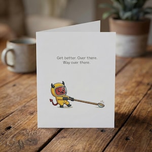 May include: A white greeting card with the text "Get better. Over there. Way over there." features a cartoon devil in a yellow hazmat suit pulling a spoon with steam rising from it. The card is on a wooden surface.