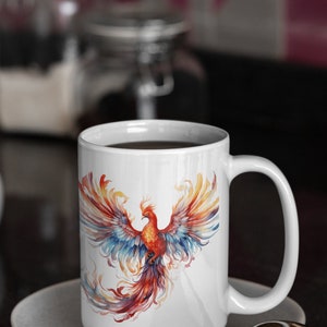 May include: White ceramic coffee mug featuring a colourful phoenix design. The phoenix is depicted with red, orange, yellow, blue, and purple feathers. The mug is filled with coffee and resting on a white saucer.