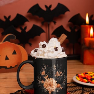 May include: Black coffee mug with a floral design featuring pumpkins, leaves, and flowers in shades of orange, black, and white. The mug has whipped cream and chocolate chips on top and a cinnamon stick sticking out.
