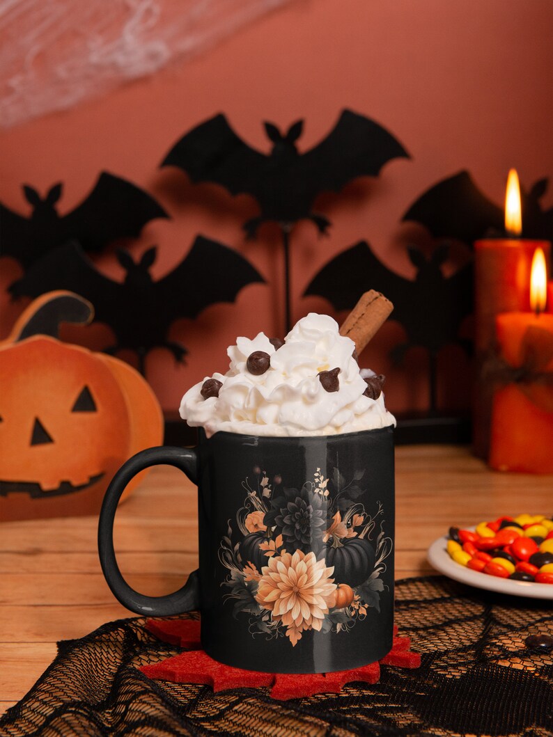 May include: Black coffee mug with a floral design featuring pumpkins, leaves, and flowers in shades of orange, black, and white. The mug has whipped cream and chocolate chips on top and a cinnamon stick sticking out.