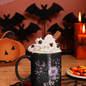 May include: Black coffee mug with a purple floral design and a white whipped cream topping. The mug is sitting on a black lace doily with a red felt coaster. There are candy corn and bats in the background.