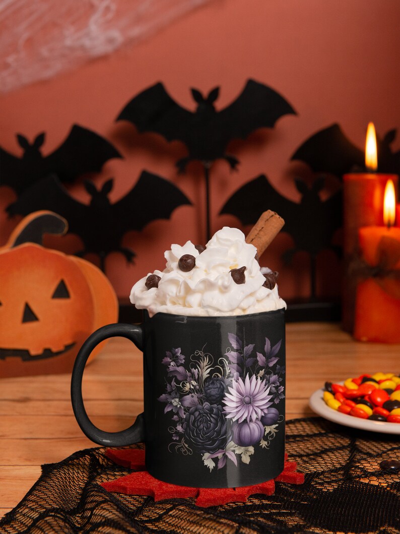 May include: Black coffee mug with a purple floral design and a white whipped cream topping. The mug is sitting on a black lace doily with a red felt coaster. There are candy corn and bats in the background.