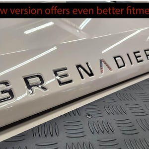 May include: A close-up of a white car with a black "GRENADIER" emblem on the back. The emblem has a small American flag design incorporated into the "A".
