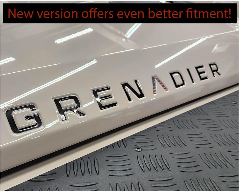 May include: A close-up of a white car with a black "GRENADIER" emblem on the back. The emblem has a small American flag design incorporated into the "A".
