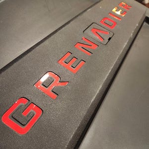 Puede incluir: Primer plano de un objeto negro con la palabra "GRENADIER" en letras rojas. Las letras están delineadas en rojo y parecen estar en relieve sobre la superficie. El objeto tiene una superficie texturizada.