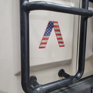 May include: A black metal ladder rack with a red, white, and blue American flag decal in the shape of a letter A.
