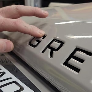 May include: Close-up of a gray car with black lettering that reads "GRE" on the back. The lettering is raised and has a glossy finish.