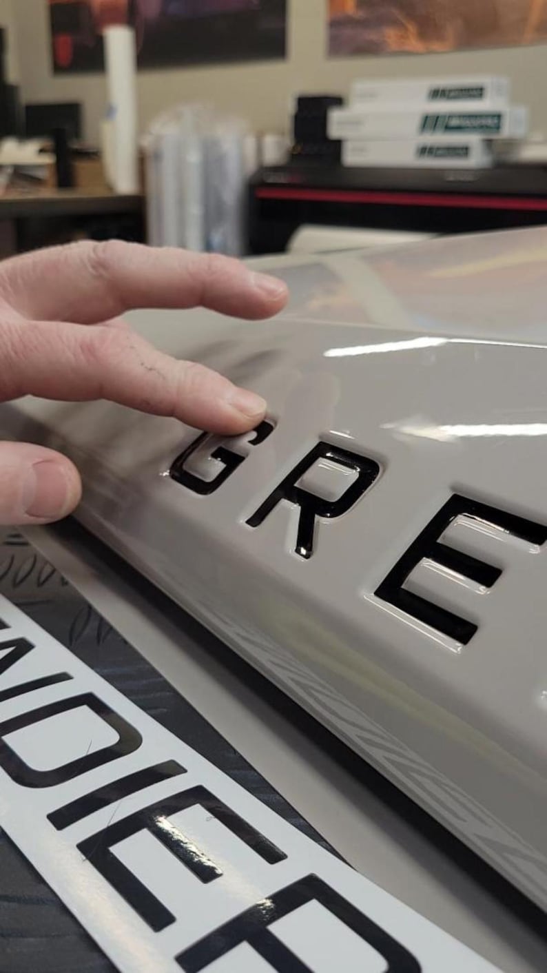 May include: Close-up of a gray car with black lettering that reads "GRE" on the back. The lettering is raised and has a glossy finish.