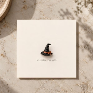 May include: A square off-white greeting card featuring a small, three-dimensional black witch hat with an orange ribbon band. The text "witching you well" is printed in a typewriter-style font in black ink below the hat on the card surface.
