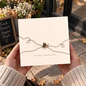 May include: A square off-white greeting card featuring two miniature figures sitting together on a thin dark green thread that forms swirling patterns. The text "best thing i accidentally found." is printed in black typewriter font below the figures.