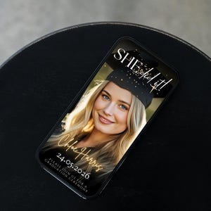 May include: A smartphone displays a graduation announcement. The screen shows a smiling blonde woman in a graduation cap with the text "SHE did it!" and "Chloe Harris 24.05.2026". The phone rests on a black surface.