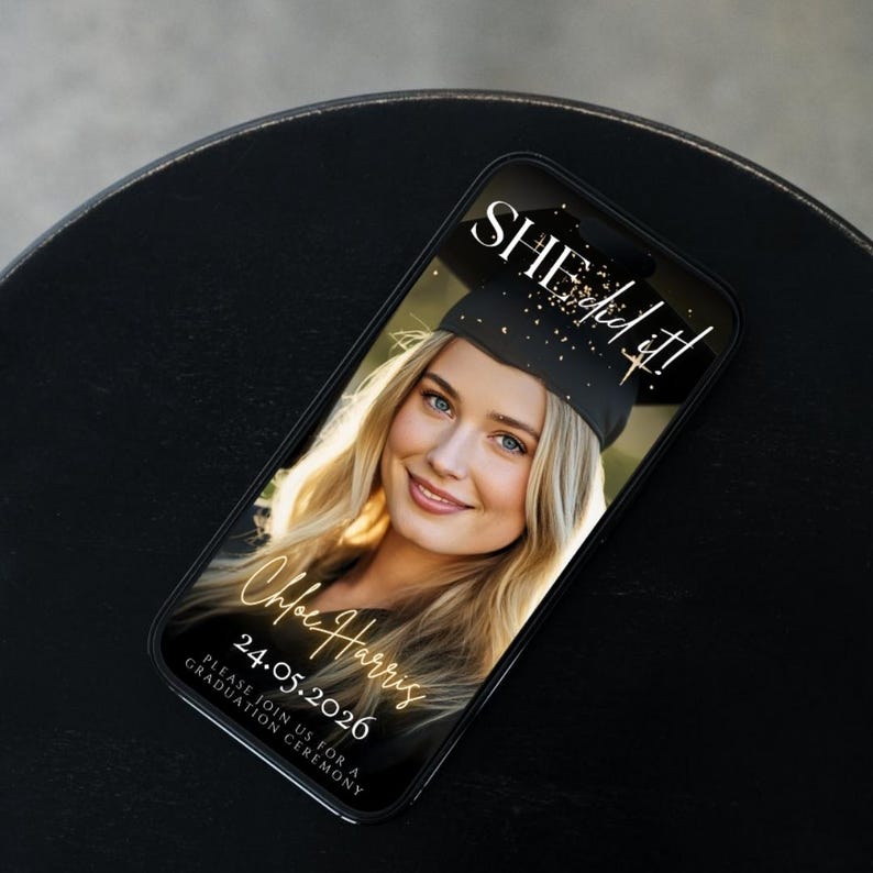 May include: A smartphone displays a graduation announcement. The screen shows a smiling blonde woman in a graduation cap with the text "SHE did it!" and "Chloe Harris 24.05.2026". The phone rests on a black surface.