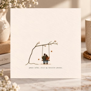 May include: A square greeting card with a cream-colored background featuring a small illustration of two figures sitting on a swing hanging from a tree branch. The text below the illustration reads, years later. still my favorite person.