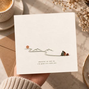 May include: A square greeting card with a textured white surface featuring a minimalist illustration of a winding path, mountains, a sun, and two figures sitting together. The text reads: wherever we end up, i'm glad it's with you.