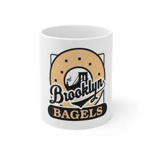 May include: White ceramic mug with a black and brown graphic of a bagel with the Brooklyn Bridge in the center and the text "Brooklyn Bagels" underneath.