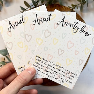 May include: Three white cards with the words "Anxiety Bear" and "Anxiet" in a handwritten font. The cards have a pattern of pastel hearts and text that reads "When things feel a bit little scary here's what to do..." The cards are held in a hand.