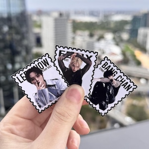 May include: Three Stray Kids stickers held in a hand. Each sticker features a different member: Lee Know, Hyunjin, and Bang Chan. The stickers are rectangular with a postage stamp border and text that reads "Stray Kids".