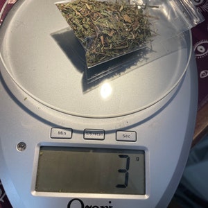 May include: A digital kitchen scale with a white and silver design displays "3" grams. A small plastic bag containing dried green herbs sits on the scale.