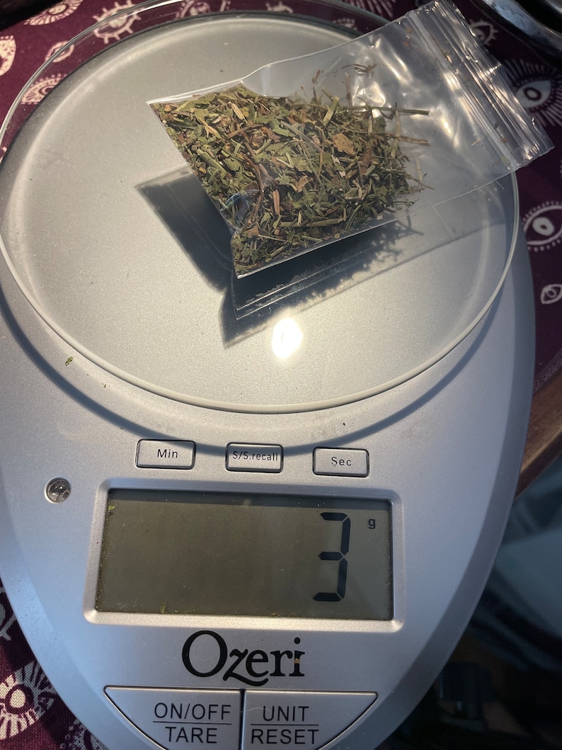 May include: A digital kitchen scale with a white and silver design displays "3" grams. A small plastic bag containing dried green herbs sits on the scale.