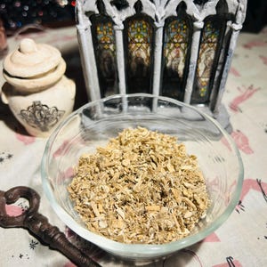 May include: A small glass bowl filled with dried, light brown, shredded herbs. The bowl is sitting on a white and pink patterned fabric.
