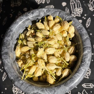 May include: A close-up of a gray marble bowl filled with dried white jasmine flower buds. The bowl is sitting on a black background with a white crystal pattern.