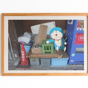 May include: A framed photograph of a blue cartoon character standing on a wooden crate in front of a cluttered background. The character has a red collar with a yellow bell and is holding a green crate. The background includes a red and white traffic cone, a blue vending machine, and various other items.