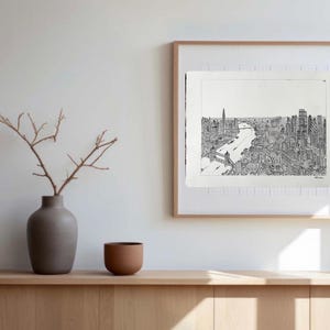 May include: A framed black and white cityscape print hangs on a white wall. The artwork depicts a detailed view of a city with a river running through it. A vase with branches and a small brown bowl sit on a wooden cabinet.