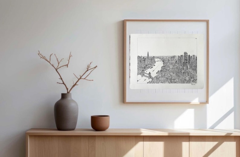 May include: A framed black and white cityscape print hangs on a white wall. The artwork depicts a detailed view of a city with a river running through it. A vase with branches and a small brown bowl sit on a wooden cabinet.