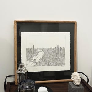 May include: A framed black and white cityscape print with a wooden frame. A cocktail shaker, a glass jar, a small white container, and a small sculpture of a head are on a dark wooden bar cart.