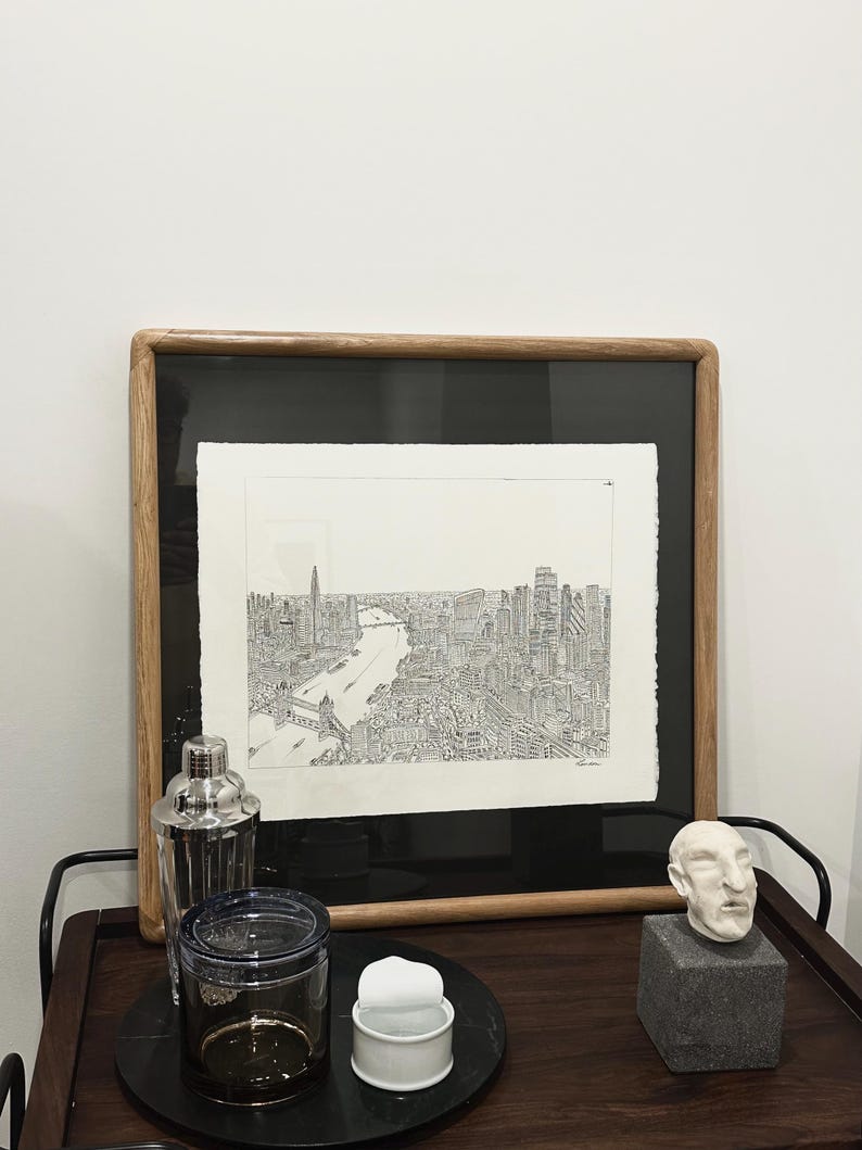 May include: A framed black and white cityscape print with a wooden frame. A cocktail shaker, a glass jar, a small white container, and a small sculpture of a head are on a dark wooden bar cart.