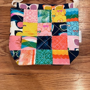May include: A patchwork tote bag with a navy blue top featuring a sunglasses pattern. The body of the bag is a quilt of colorful squares with tropical and sunglasses designs. It has a white drawstring handle.