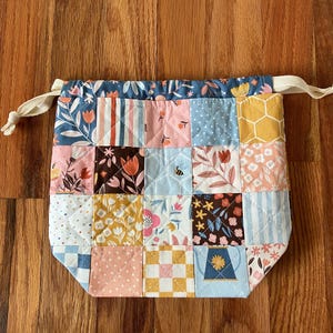 May include: A patchwork drawstring bag with a floral and geometric pattern. The bag features a variety of fabrics in shades of pink, blue, yellow, and brown. The top of the bag is a dark blue floral print, and it has a cream-colored drawstring.