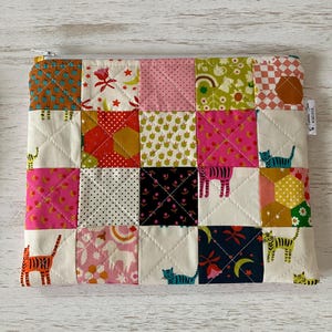May include: A patchwork zippered pouch with a variety of colorful fabric squares. The squares feature patterns like cats, stars, and floral designs in shades of pink, orange, green, and black. The pouch is quilted with white thread.