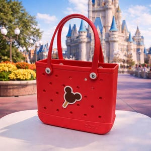 Puede incluir: Un bolso tote rojo brillante con una pegatina de helado de Mickey Mouse. El bolso tiene dos asas y peque&ntilde;os agujeros. El fondo presenta un castillo y un d&iacute;a soleado.