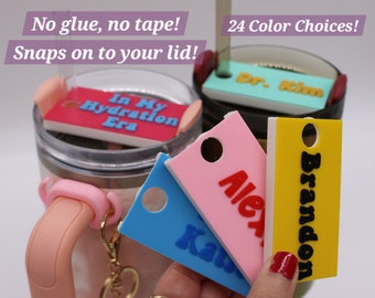 May include: A set of colorful, personalized drink lid tags with names "Brandon", "Alex", and "Ka" printed in black. The tags are made of a durable material and have a hole for a straw. The tags are shown with a pink, blue, and yellow background. The text "24 Color Choices!" is visible in the background.