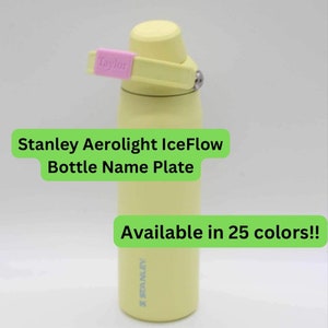 Puede incluir: Una botella de agua amarilla Stanley Aerolight IceFlow con una etiqueta de nombre rosa que dice "Taylor". La botella tiene una tapa negra y un logotipo azul de Stanley en la parte inferior. El texto "Stanley Aerolight IceFlow Bottle Name Plate" está escrito en negro sobre fondo verde. El texto "Disponible en 25 colores!" está escrito en negro sobre fondo verde.