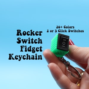 Könnte beinhalten: Ein grüner Fidget-Schlüsselanhänger mit Wippschalter und schwarzem Schalter. Der Schlüsselanhänger hat einen silbernen Ring und wird vor einem hellblauen Hintergrund gehalten. Der Text links lautet "Rocker Switch Fidget Keychain". Der Text oben rechts lautet "20+ Colors 2 or 3 Click Switches".