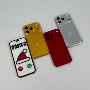 以下が含まれることがあります： 金、赤、銀色の4つの装飾的な携帯電話ケース。1つのケースにはサンタの帽子と画面に「サンタ」の文字が表示されています。他のケースにはカメラアレイとAppleのロゴがあります。