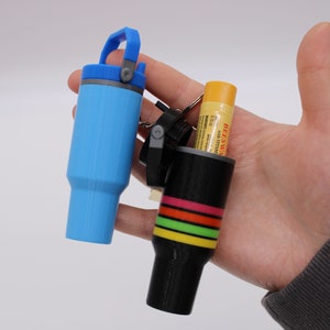 Może przedstawiać: Dwa małe, drukowane w 3D pojemniki w kształcie kubka. Jeden jest jasnoniebieski z pasującym uchwytem i pokrywką. Drugi jest czarny z poziomymi paskami w kolorze różowym, pomarańczowym, żółtym i zielonym. W czarnym pojemniku widoczna jest tubka balsamu do ust.