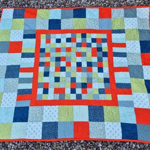 K&ouml;nnte beinhalten: Ein farbenfroher Patchwork-Quilt mit quadratischem Muster. Der Quilt zeigt Quadrate in Blau-, Gr&uuml;n-, Orange- und Wei&szlig;t&ouml;nen. Der Quilt hat einen orangefarbenen Rand und ein zentrales quadratisches Design. Der Quilt ist aus Stoff.