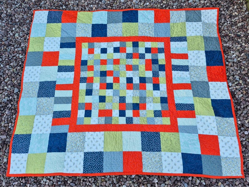 K&ouml;nnte beinhalten: Ein farbenfroher Patchwork-Quilt mit quadratischem Muster. Der Quilt zeigt Quadrate in Blau-, Gr&uuml;n-, Orange- und Wei&szlig;t&ouml;nen. Der Quilt hat einen orangefarbenen Rand und ein zentrales quadratisches Design. Der Quilt ist aus Stoff.