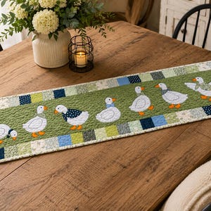 May include: A rectangular quilted table runner featuring a central olive green panel with six white goose appliqu&eacute;s wearing patterned scarves. The border consists of a patchwork of small squares in shades of blue, green, and white floral prints.