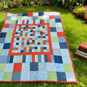 K&ouml;nnte beinhalten: Ein bunter Patchwork-Quilt mit rotem Rand und einem quadratischen Mitteldesign. Der Quilt zeigt Quadrate aus blauen, gr&uuml;nen und roten Stoffen. Der Quilt liegt auf einer Rasenfl&auml;che, mit einem Stapel B&uuml;cher in der N&auml;he.