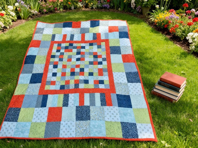 K&ouml;nnte beinhalten: Ein bunter Patchwork-Quilt mit rotem Rand und einem quadratischen Mitteldesign. Der Quilt zeigt Quadrate aus blauen, gr&uuml;nen und roten Stoffen. Der Quilt liegt auf einer Rasenfl&auml;che, mit einem Stapel B&uuml;cher in der N&auml;he.